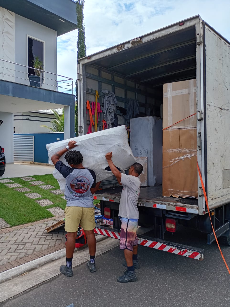 Dois ajudantes de uniforme erguendo um colchão embalado pra dentro do caminhão baú, na frente de uma casa