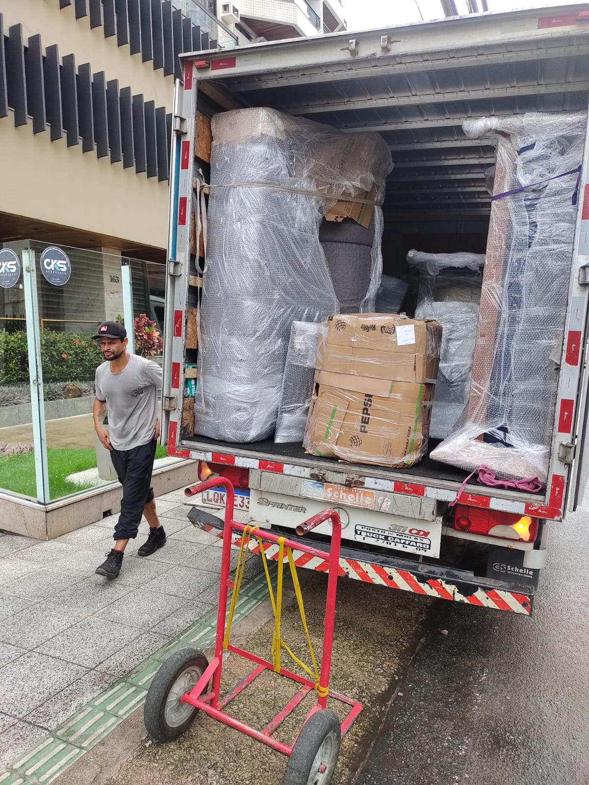 Baú da Sprinter carregado com sofá e móveis embrulhados em plástico-bolha, ajudante ao lado e carrinho de carga
